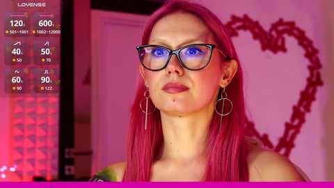 A video of a fair-skinned woman with long, straight pink hair and blue eyes, wearing black-framed glasses and hoop earrings. She has a heart-shaped outline on a pink wall behind her.