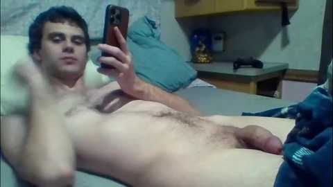 A video of a naked young man with short dark hair, holding a smartphone, lying on a bed in a bedroom.