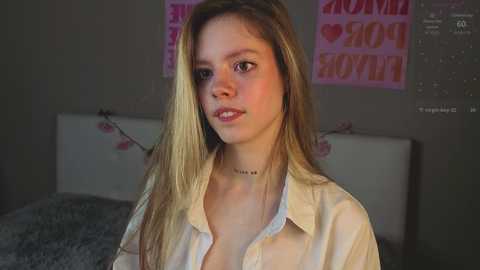 Video of a young Caucasian woman with long, straight blonde hair, wearing a partially unbuttoned white shirt, in a dimly lit bedroom with pink motivational posters.