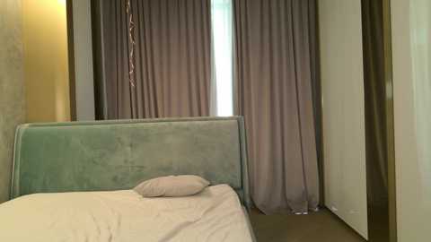 Video of a minimalist bedroom featuring a pale green velvet headboard, white bed sheets, a single pillow, and lavender curtains. The room has beige walls and a wooden door frame.