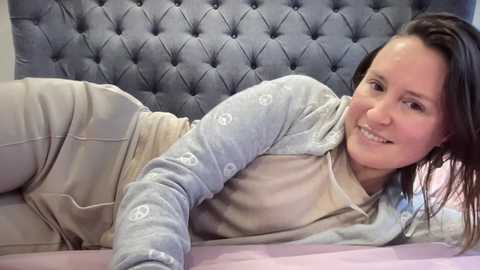 Video of a smiling woman with fair skin and brown hair lying on a bed with a tufted, gray headboard, wearing light gray pajamas with white peace signs.