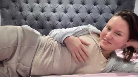 Video of a fair-skinned, brunette woman with medium build, wearing a light gray sweater and beige pants, lying on a bed with a tufted gray headboard, resting her head on her hand.