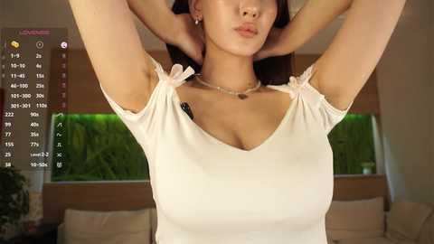 A video of an Asian woman with medium skin tone, long black hair, and a medium-sized bust, wearing a beige sleeveless top with bows. She's in a modern living room with green foliage outside the window.