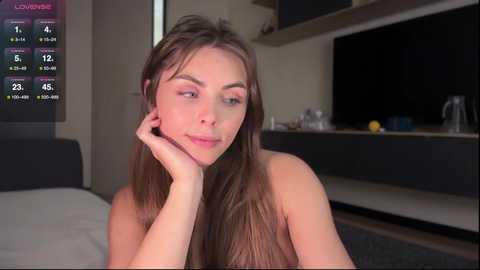 Video of a young, fair-skinned woman with long brown hair, resting her chin on her hand, in a modern bedroom with a flat-screen TV and digital clock displaying 4:23.