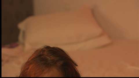 Video of a woman's head and shoulder, lying on a beige bed with blurred pillows and bedding, her dark hair slightly messy, and a warm, cozy ambiance.