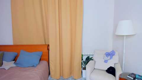 Video of a minimalist bedroom with a beige curtain, orange headboard, blue pillows, a white armchair, a tall floor lamp, and a cluttered nightstand.