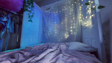 A video of a cozy bedroom with a bed covered in a soft, light pink blanket, fairy lights draped over the white walls, and green plants hanging from the ceiling.