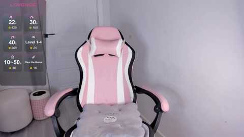 A video of a pink gaming chair with white accents, situated in a minimalist room with white walls, a roll of toilet paper, and a screen displaying a video game interface.