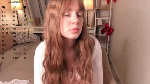 Video of a young Caucasian woman with long, wavy, light brown hair, wearing a white sweater, sitting in a cozy room with beige walls and a white lamp.