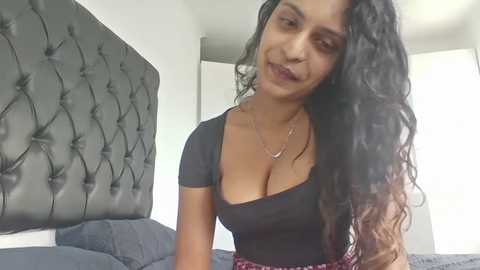 Video of a South Asian woman with long, wavy black hair, wearing a black, low-cut top, smiling in a modern, minimalist bedroom with a tufted gray headboard.