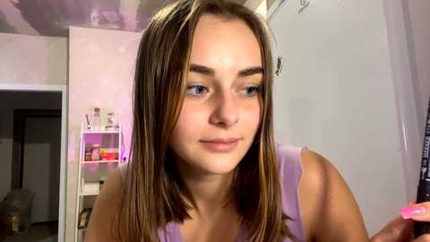 Video of a young woman with fair skin, straight brown hair, and light makeup, wearing a lavender sleeveless top. She is indoors in a brightly lit room with a white wall and open door.