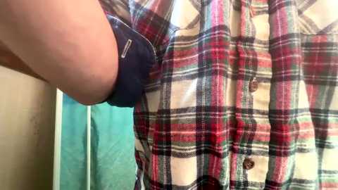 A close-up video shows a person's arm in a dark blue shirt, with a colorful plaid jacket buttoned up. The background includes a turquoise wall and part of a beige cabinet. The image is casual and indoor.