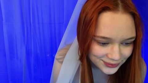 Video of a fair-skinned woman with long, straight, reddish-brown hair, wearing a white top, smiling, against a vibrant blue backdrop.