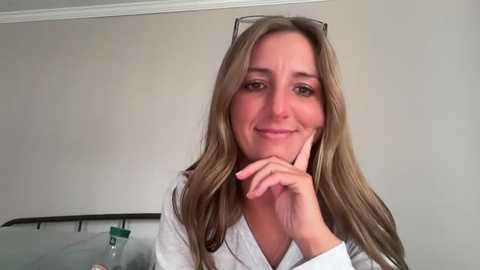 Video of a Caucasian woman with long, wavy blonde hair, wearing glasses, smiling. She is dressed in a white robe, leaning on a bed with a light beige wall behind her.