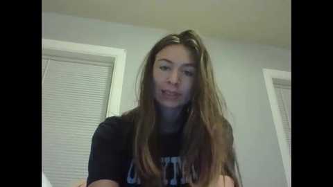 A video of a young woman with long, light brown hair, wearing a black t-shirt, indoors with light gray walls and closed blinds in the background.