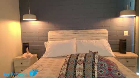 Video of a cozy bedroom with a wooden headboard, white bedding, colorful throw pillows, and two side tables with black lamps and beige chairs. The background features a textured gray brick wall.