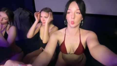 Media: Video of three women in a dimly lit room. They are in various states of undress, with one woman in a red bikini, another in a black bra, and the third in a black tank top. They are posing and making faces, with one woman taking a selfie.