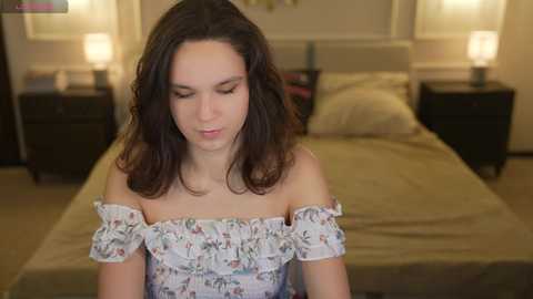 Video of a young woman with long, dark hair, wearing a floral off-shoulder top, sitting in a dimly lit bedroom with a neatly made bed, two bedside lamps, and beige walls.