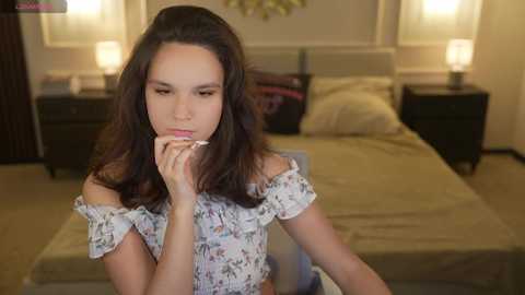 Video of a young woman with medium-length dark hair, wearing a floral off-shoulder dress, sitting pensively in a softly lit bedroom with beige walls, a bed, and two nightstands.