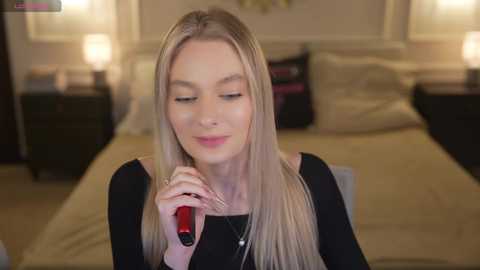 Media: Video of a blonde woman with fair skin, wearing a black top, applying red lipstick in a cozy bedroom with beige furniture and soft lighting.