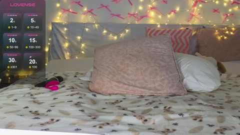 A video of a cozy bedroom with a bed covered in a floral-patterned duvet, featuring a pink pillow and white pillows. The background is adorned with fairy lights and pink ribbons, and a smartphone displaying a game screen is visible on the left.
