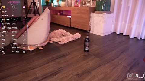 Video of a messy bedroom with a dark wooden floor, a stuffed pink bear, a bottle of wine, and a messy bed.