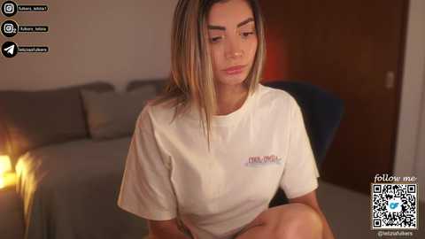 Video of a young woman with straight, shoulder-length blonde hair, wearing a white T-shirt and sitting on a couch in a dimly lit living room.