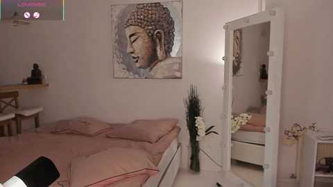 Media: Video of a minimalist bedroom with a white bed, pink bedding, Buddha wall art, white-framed mirror, and a vase with white flowers.