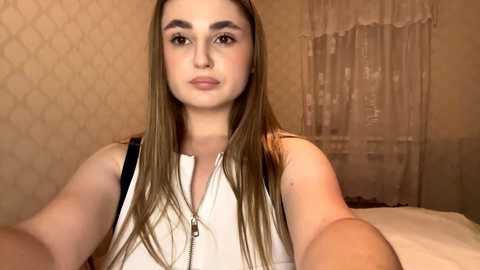 Video of a young Caucasian woman with long, straight brown hair, wearing a white sleeveless top, in a beige room with patterned wallpaper and a semi-transparent curtain.
