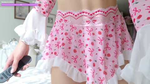 Video of a person wearing a pink, floral-patterned ruffled romper with white lace trim, holding a remote. Background shows a bedroom with white bedding, framed pictures, and a TV.