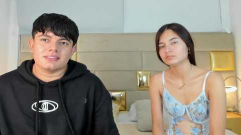 Video of a young man with short black hair in a black hoodie and a young woman with straight dark hair in light blue lace lingerie, standing in a modern bedroom with a white headboard and gold accents.