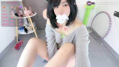 Video of an Asian woman with shoulder-length black hair, wearing a gray off-shoulder sweater and a white handkerchief gag, sitting on a tiled floor, surrounded by sex toys and a calendar in a cluttered room.