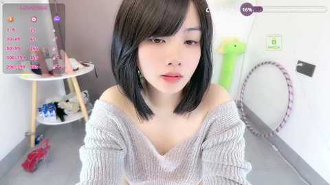 Video of a young Asian woman with straight black hair, wearing a loose grey sweater, sitting in a minimalist room with a white wall, a round table, and a green object.