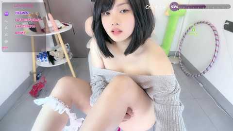 A young Asian woman with black bob haircut, fair skin, and small breasts, sits topless in a casual room. She wears a gray off-shoulder sweater and white socks.