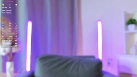 A blurred video of a cozy, dimly lit room with a gray cushioned chair in the foreground. Neon purple lights on either side create a soft glow, and a potted plant is visible on the right.