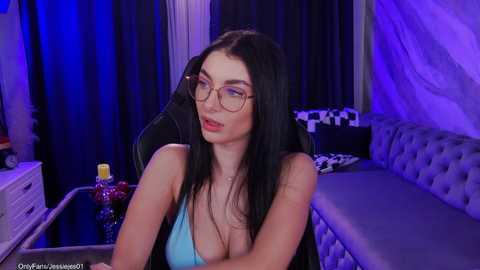 A video of a young woman with long black hair, fair skin, and glasses, wearing a low-cut blue tank top, sitting in a dimly lit room with a checkered pillow and dark curtains in the background.