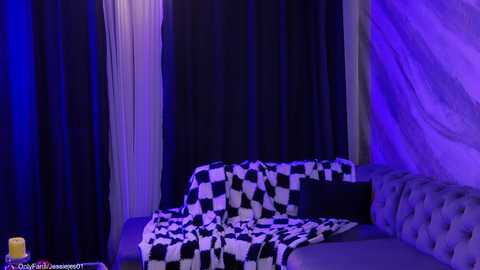 A video of a dimly lit, modern room with a checkered blanket on a tufted gray couch against dark curtains and purple lighting.