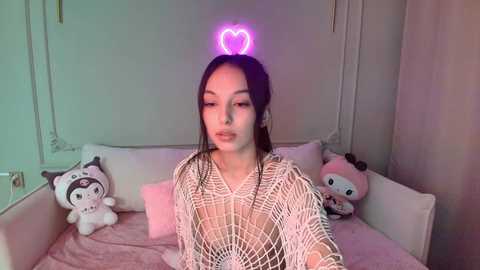 Video of a young woman with long black hair and fair skin, wearing a white fishnet top, sitting on a pink bed with plush toys, illuminated by a pink heart-shaped neon light.