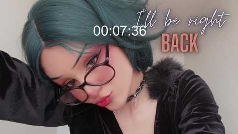 Video of a fair-skinned woman with teal hair, wearing glasses and a black velvet jacket. She has a choker necklace and a sad expression. Text overlay reads, \"I'll be right back.\
