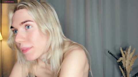 Video of a fair-skinned, blonde woman with blue eyes and light makeup, wearing a strapless top, sitting indoors. Background features beige curtains and a wooden lamp.