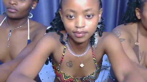 Video of three young Black women with braided hair, wearing colorful, patterned tops, against a blue curtain backdrop.