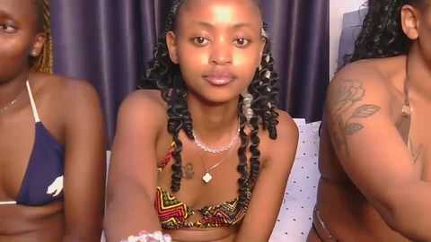 Video of three African women with braided hair, two wearing colorful bikinis, one topless, in front of purple curtains.