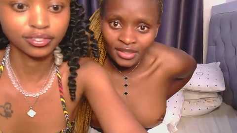 Video of two topless young Black women with braided hair, smiling, wearing beaded necklaces, lying on a bed with white sheets and a purple headboard in a dimly lit room.