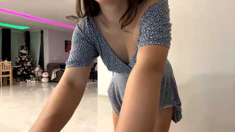 Video of a woman in a blue floral dress, leaning forward, in a modern, brightly lit living room with a Christmas tree and colorful LED strip lights.