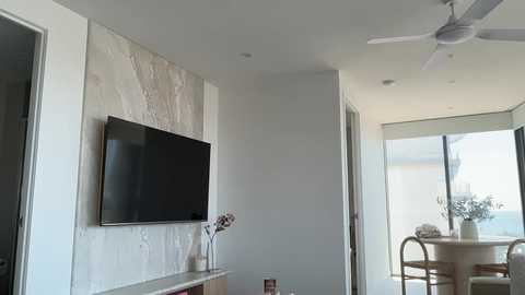 Video of a modern living room with a large flat-screen TV mounted on a textured stone wall, a small wooden table with decorative items, and a ceiling fan.