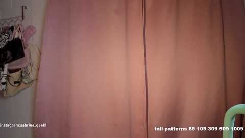 Video of a bedroom with a large, soft pink curtain covering the background, partially obscuring a white wall and a beige pillowcase with black text. The image appears to be a still from a video with a watermark.