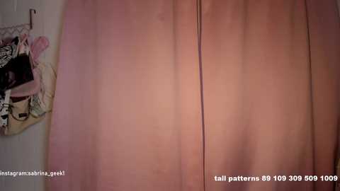 Video of a pink, pleated curtain with a slight crease, hanging in a room with a white wall. A multicolored garment is partially visible on the left.