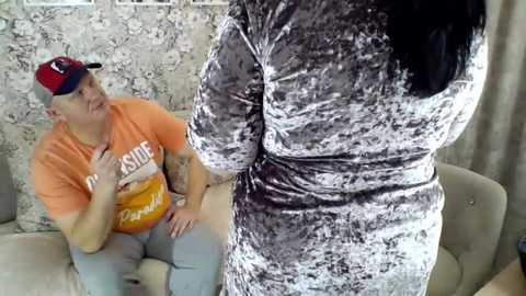 Video of a woman in a velvet dress with a man in an orange t-shirt and cap, sitting on a beige sofa in a room with floral-patterned wallpaper.