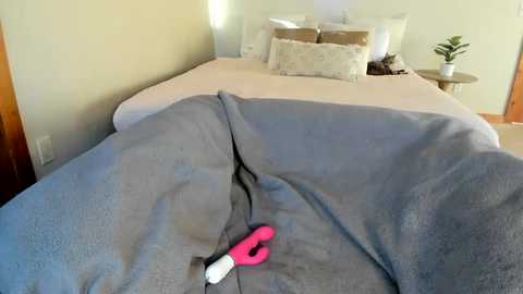 A video of a cozy bedroom with a gray blanket, a pink and white vibrator, and a neatly made bed with white pillows and a beige wall.