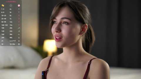 A video of a young East Asian woman with fair skin and dark hair tied back. She wears a maroon bralette and is looking to the side, smiling. The background features a blurred, dimly lit bedroom with a white bed and dark curtains.